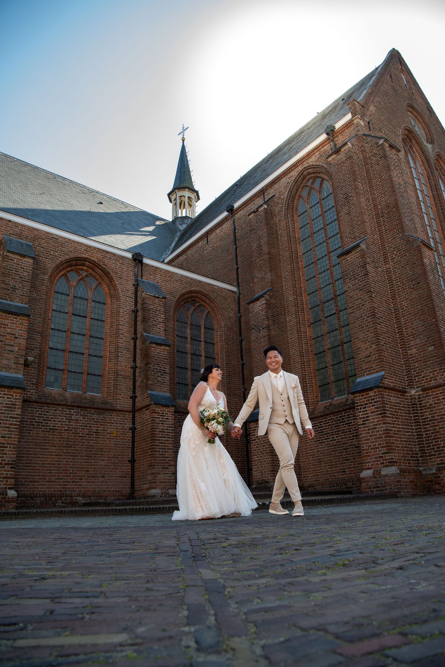 A wedding couple poses for creative angle photos outside a historic brick church with gothic windows and spires.