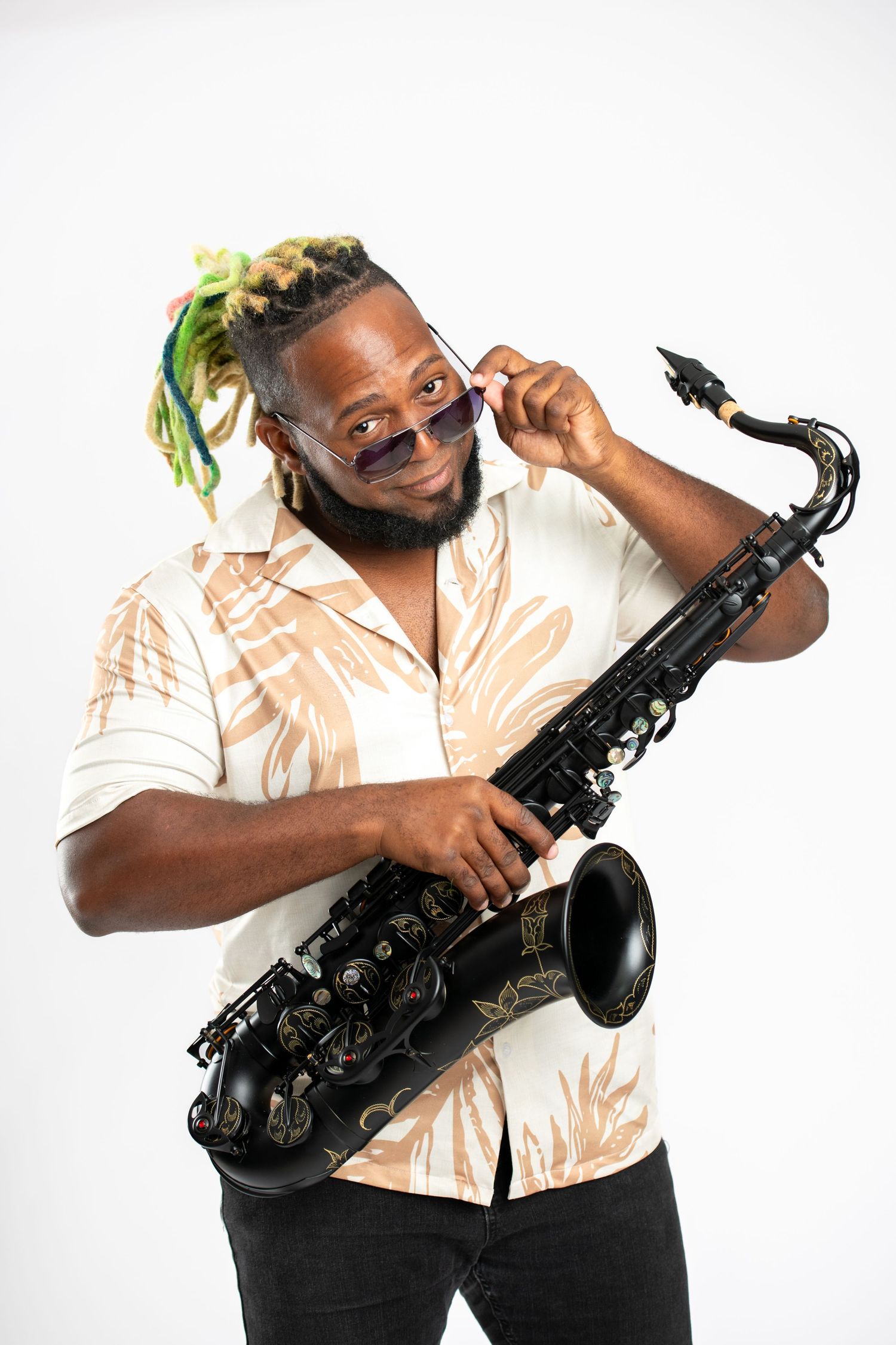 A Black musician with colorful dreadlocks holds a black saxophone while playfully lowering his sunglasses in a studio.