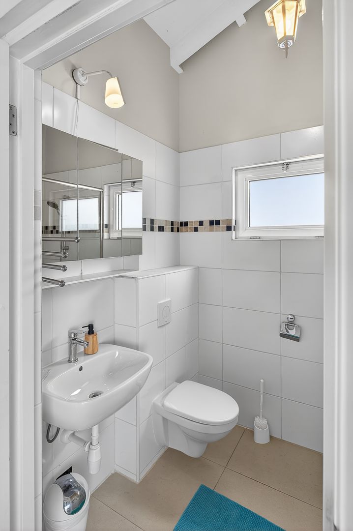 Modern white bathroom with wall-mounted toilet, pedestal sink, large mirror, mosaic tile border, and small window providing natural light.