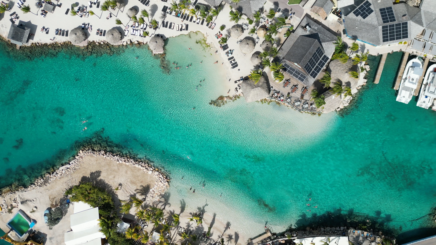 Aerial view of a tropical beach with turquoise waters, white sand, and docked boats along the shoreline.