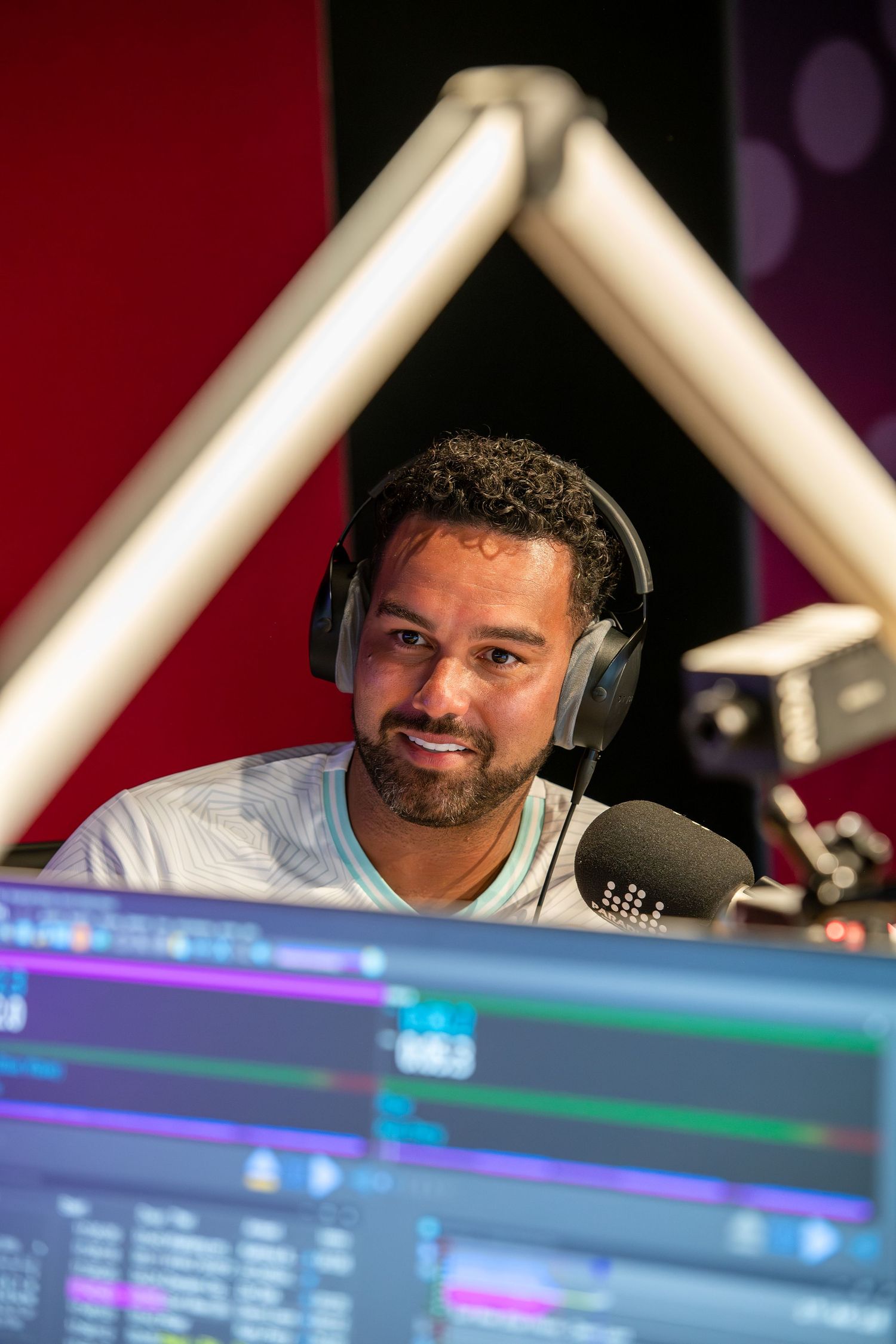Male radio host wearing headphones speaks into a microphone in a professional broadcast studio with colorful screens.