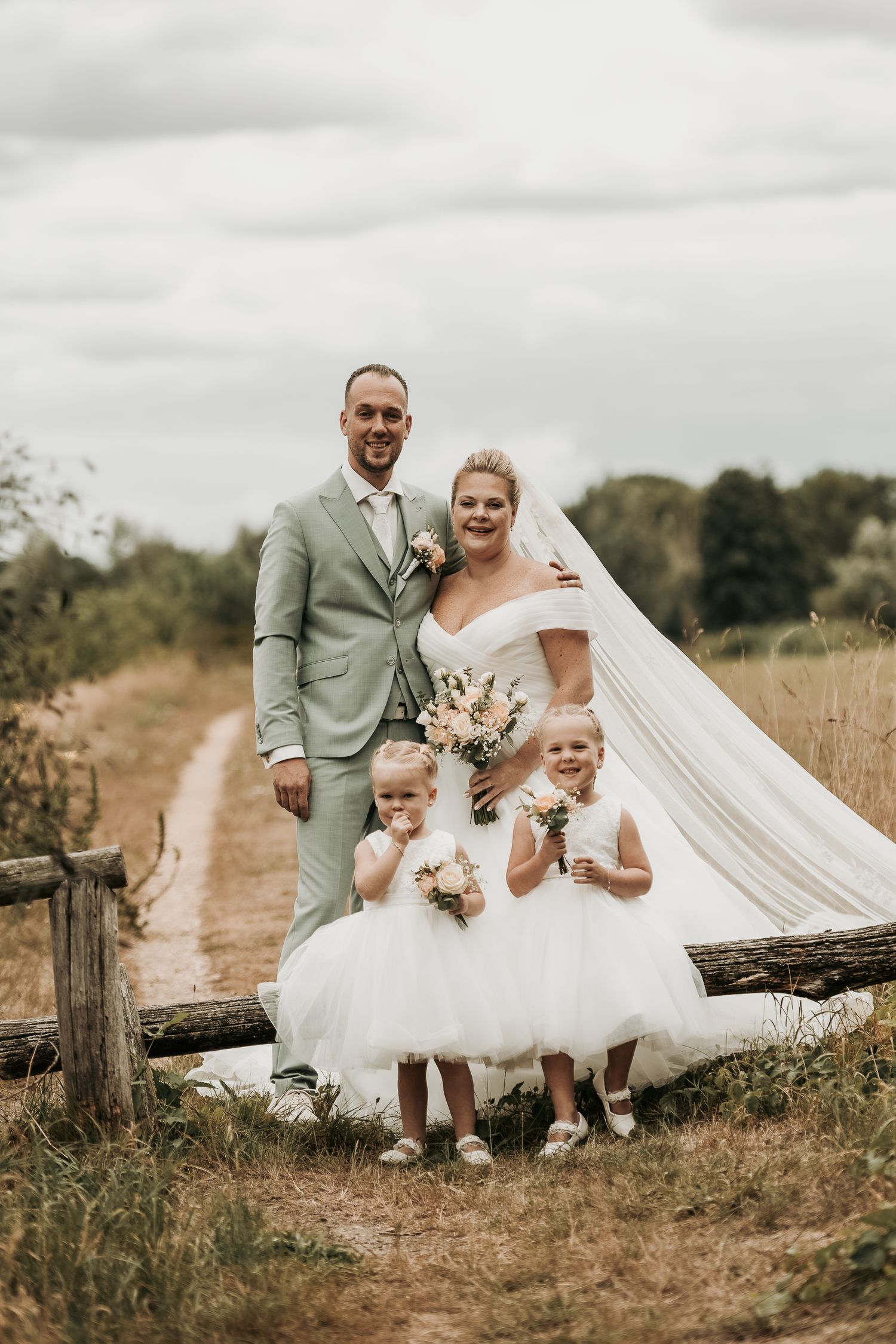 A wedding party poses together on a rustic dirt path in a grassy field on an overcast day.
