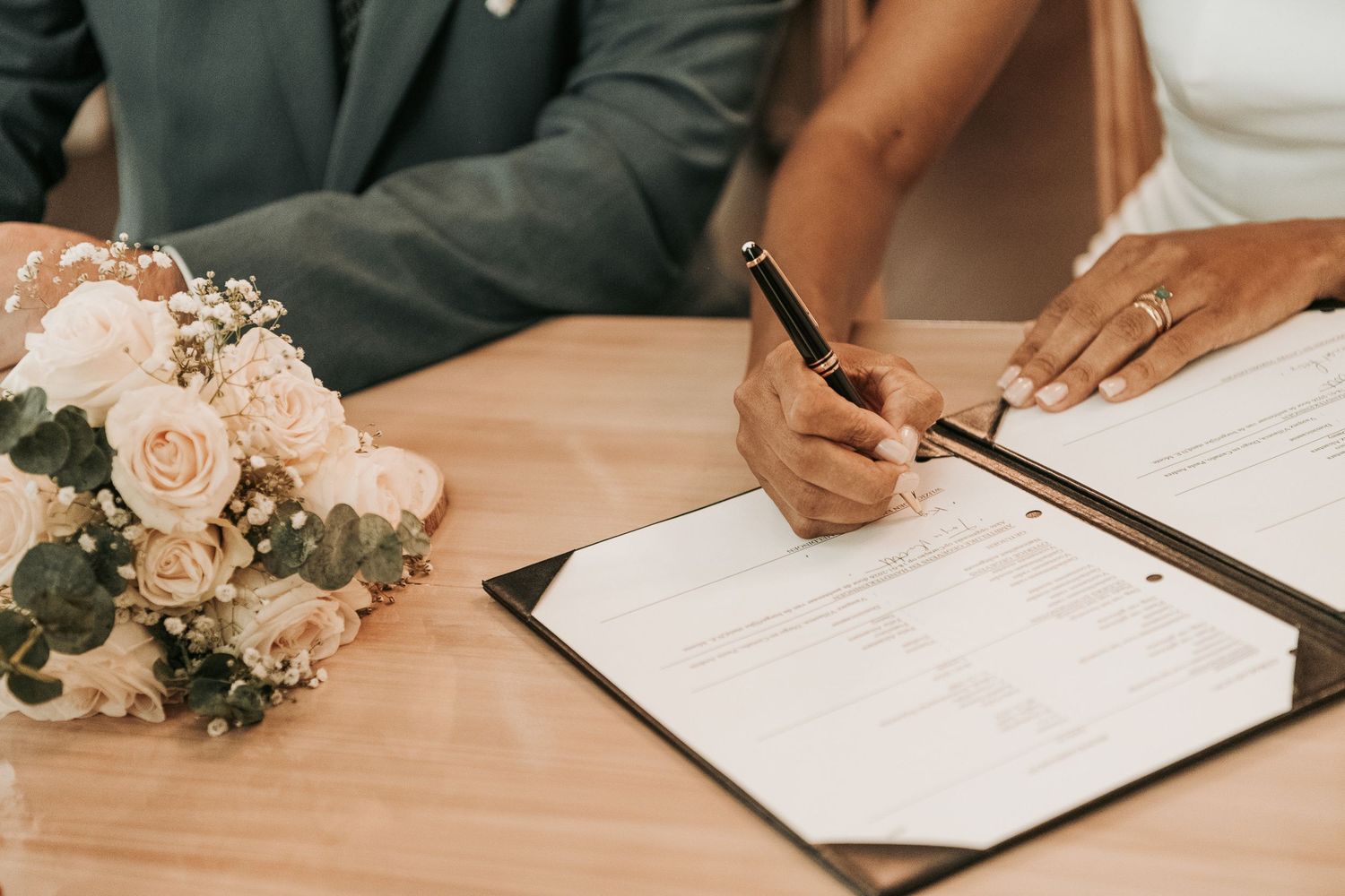 Someone signs a document next to a blush pink rose bouquet on a wooden table during a wedding ceremony.