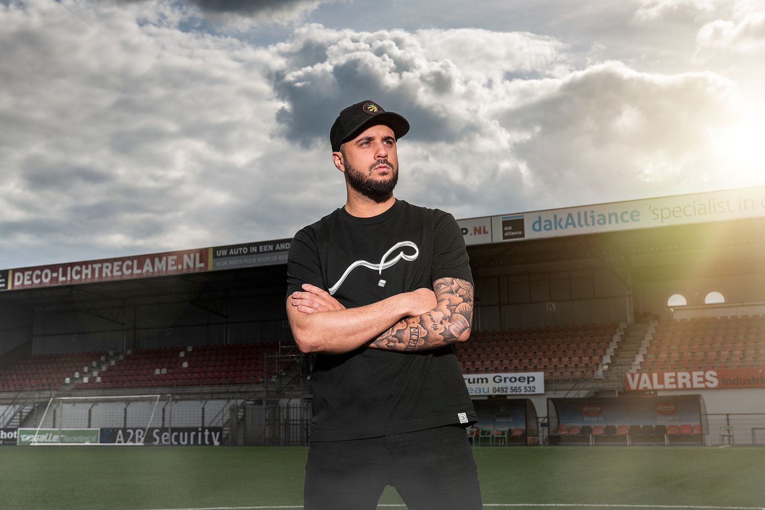 A baseball player in a black shirt stands on a field during sunset with dramatic clouds in the background.