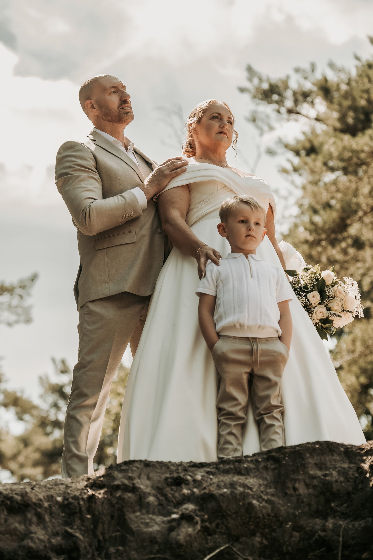 A wedding photo of a couple and young child standing together on a rocky outcrop against a cloudy sky.
