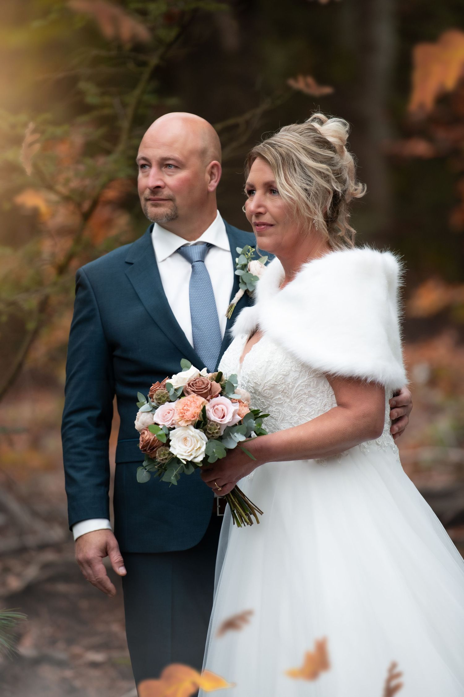 A couple in wedding attire poses outdoors in autumn, with the bride wearing a white fur wrap and holding a peach bouquet.