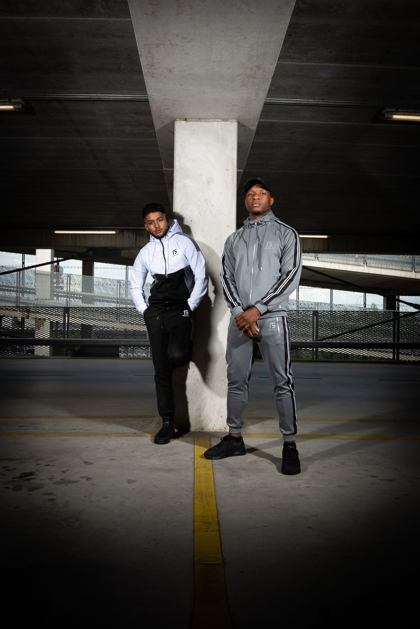 Two people in athletic wear pose dramatically under a concrete structure in a parking garage.