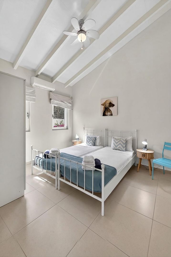 Bright white bedroom with exposed beam ceiling, metal bed frame, blue accents, ceiling fan, and tiled floors in a tropical setting.
