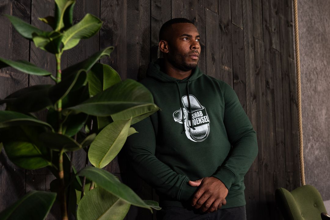 Person wearing a dark green hoodie with graphic design stands next to indoor plants against a wooden wall.