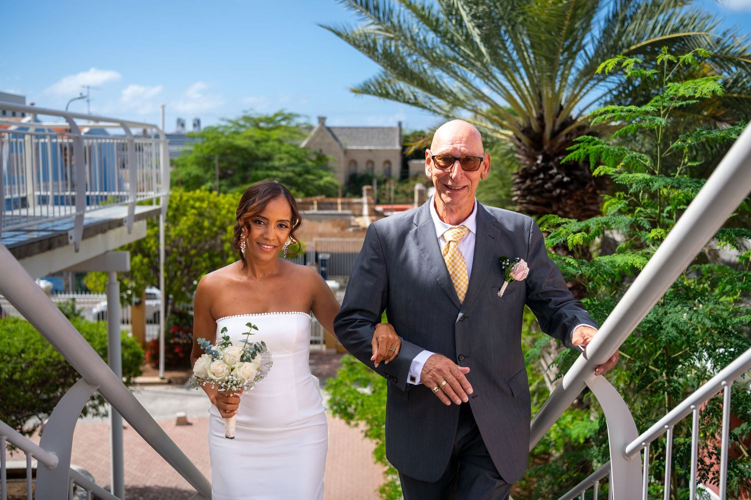 A couple walks down outdoor stairs at a tropical venue with palm trees and white flowers on a sunny day.