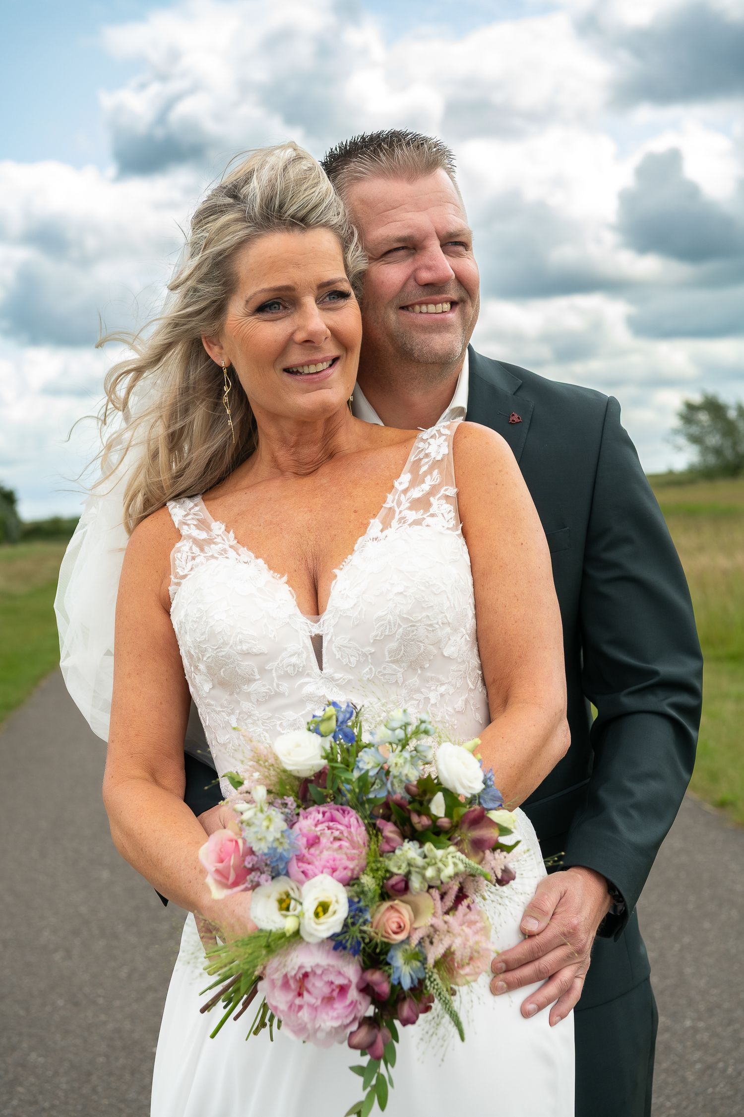 A bride in a lace wedding dress holds a pink and purple bouquet while posing outdoors with her groom on a cloudy day.