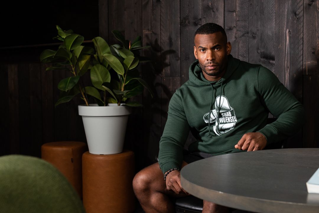 Person wearing dark green hoodie sitting at round table with houseplant in white pot nearby in dark moody setting.