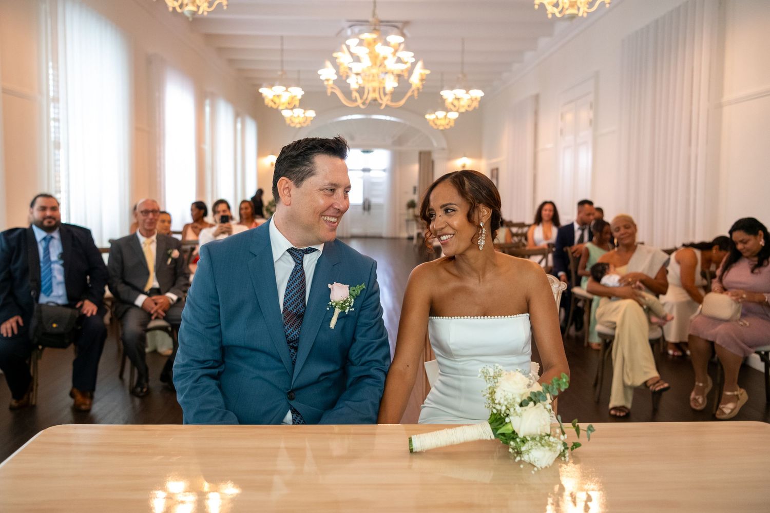 Wedding guests dance together in an elegant white ballroom with crystal chandeliers overhead during a reception celebration.