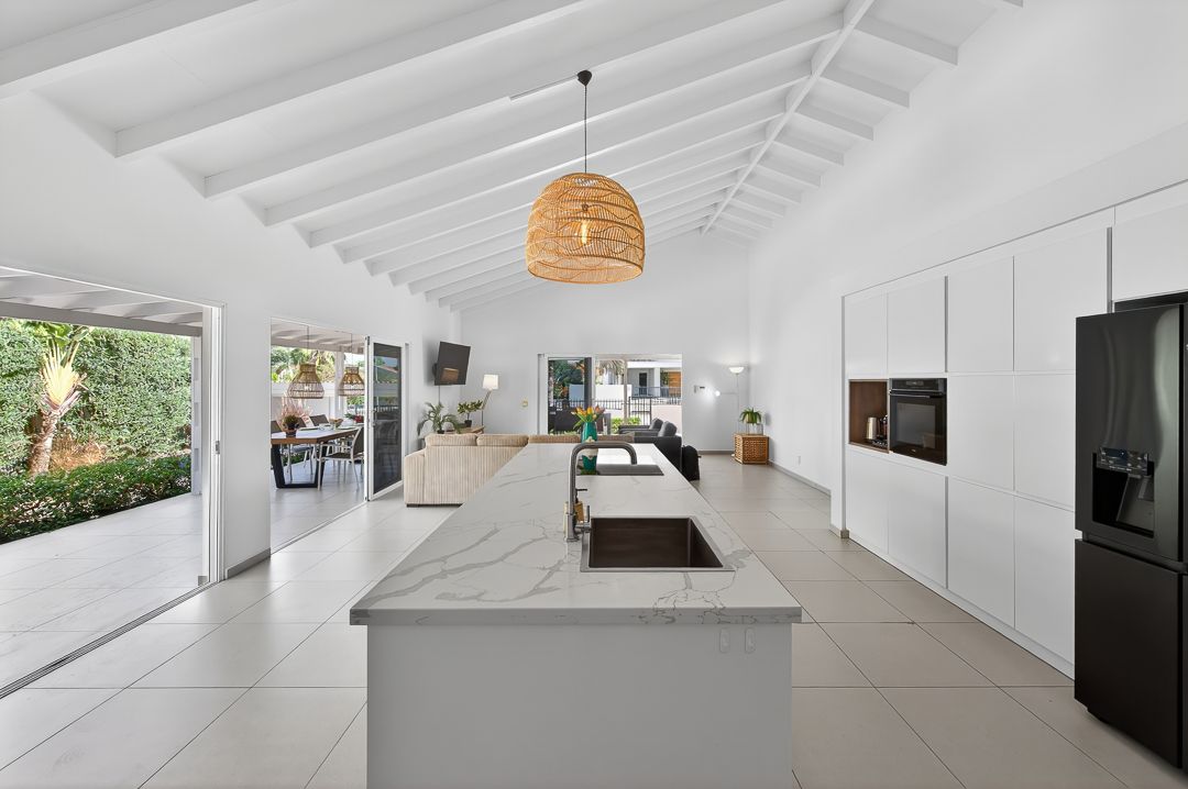 Modern open-plan kitchen with marble island, white cabinetry, rattan pendant light, and vaulted ceiling leading to outdoor area.