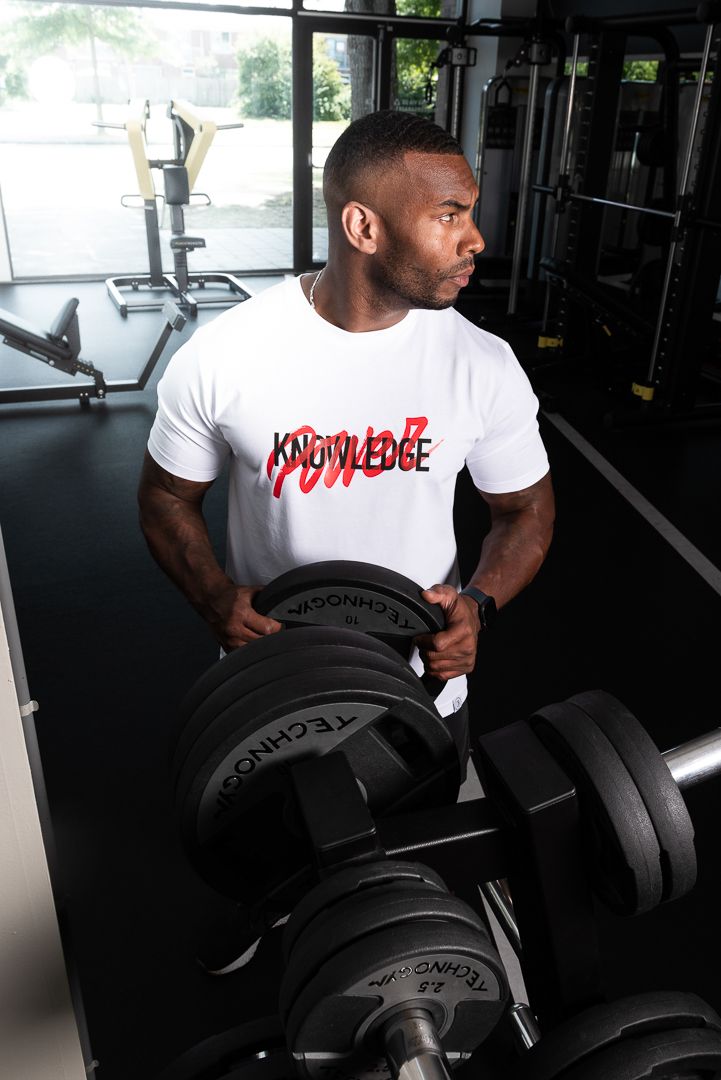 Athletic person in white 'Knowledge Power' t-shirt selecting weights from dumbbell rack in modern gym setting.