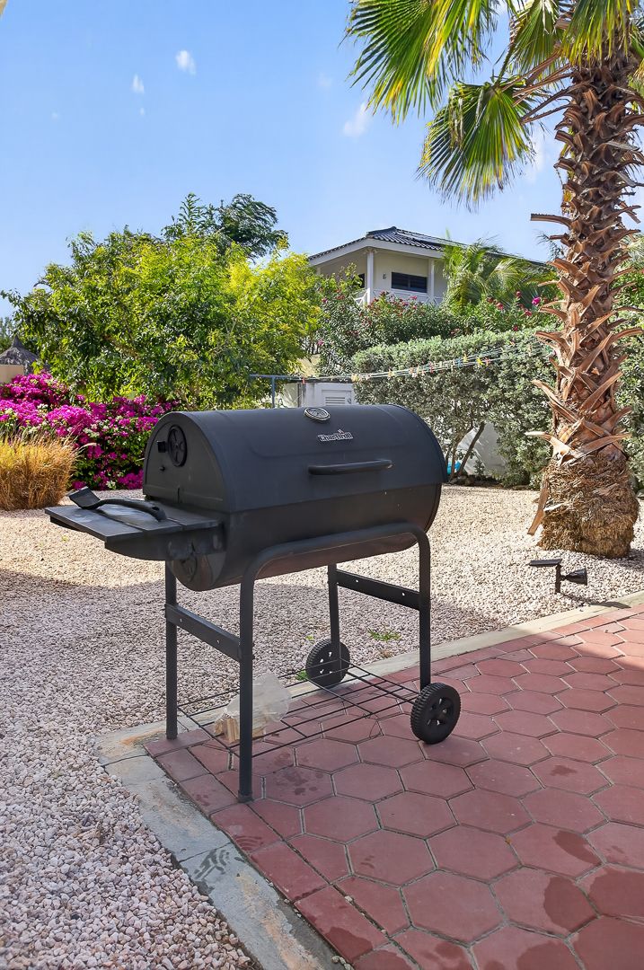 Black Char-Broil charcoal barrel grill on patio with palm trees, colorful flowers, and a house in the background.
