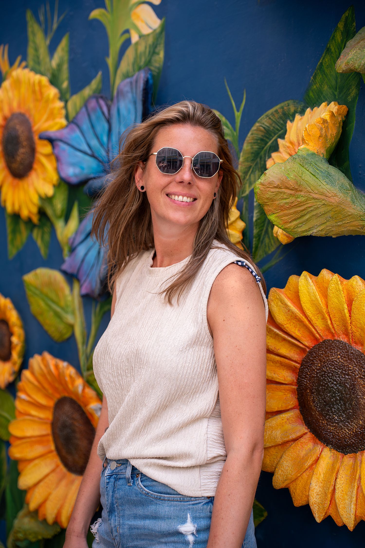 Casual summer outfit with white sleeveless top and jeans against vibrant sunflower wall backdrop with blue background.