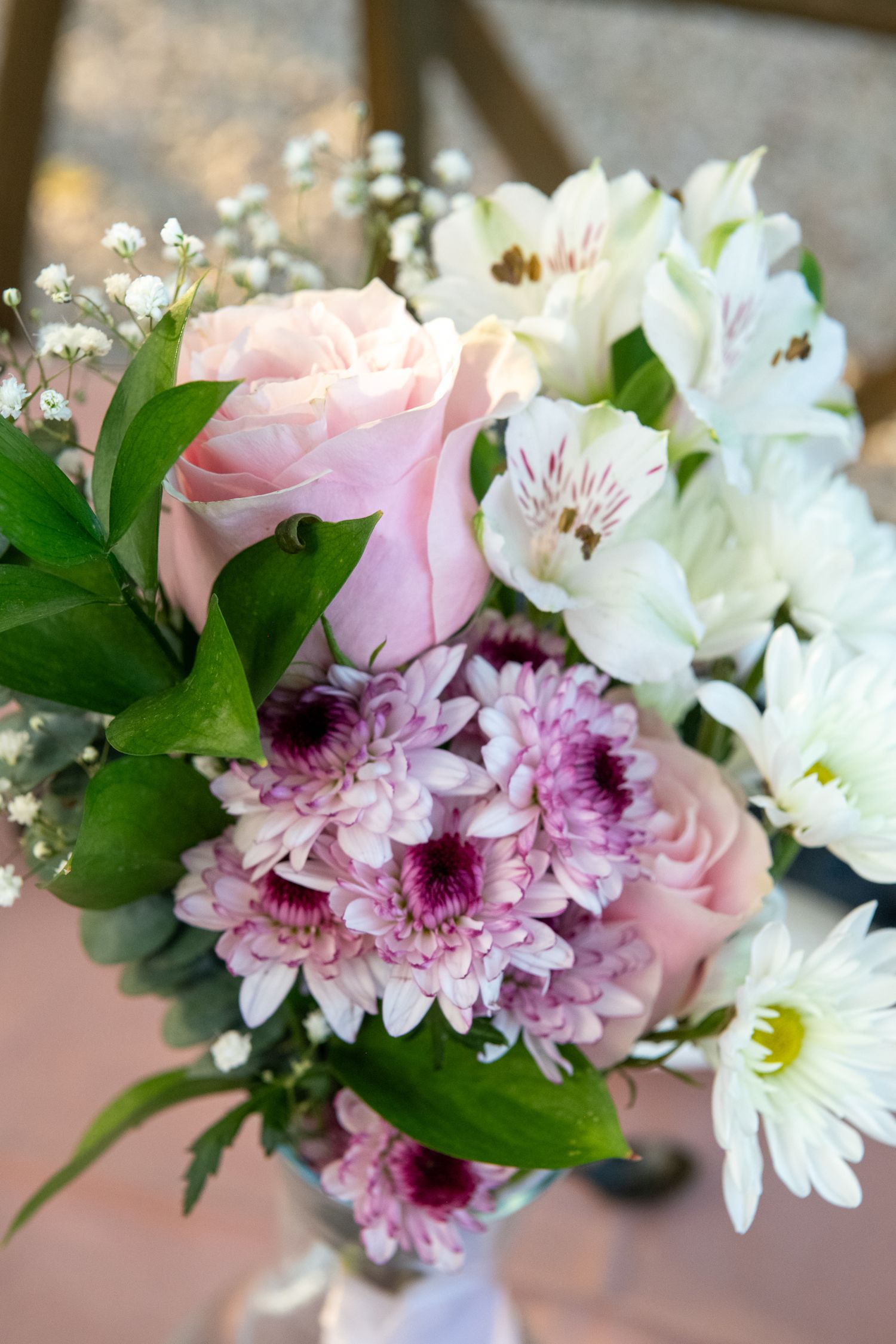 A delicate floral arrangement featuring pink roses, white daisies, purple chrysanthemums and baby's breath against a soft pink background.