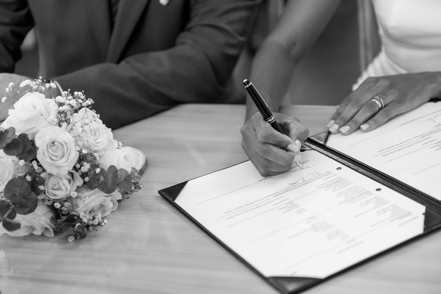A wedding bouquet of roses rests on a table while hands sign the marriage register in black and white.