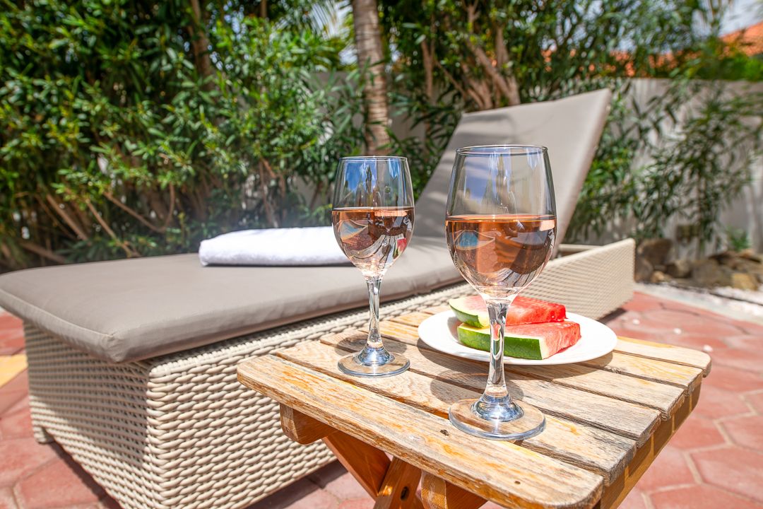 Two glasses of rosé wine with a fruit plate on a wooden table beside a lounge chair in a sunny outdoor patio.