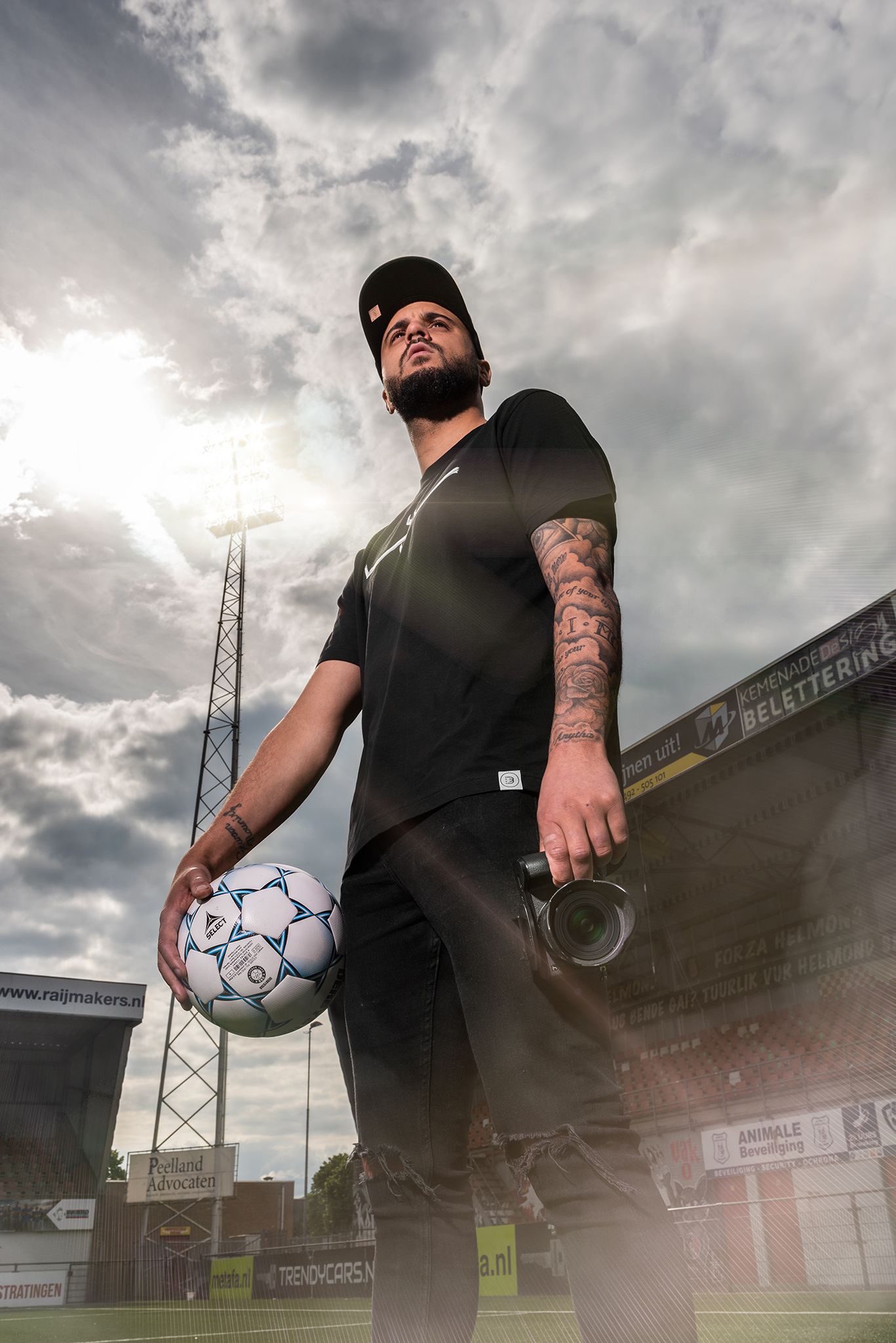 A person in black clothing performs soccer ball tricks against a dramatic cloudy sky at a sports stadium.
