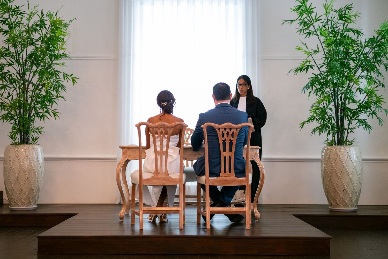 Three people sit at a wooden table in a bright room decorated with potted bamboo plants and beige walls.
