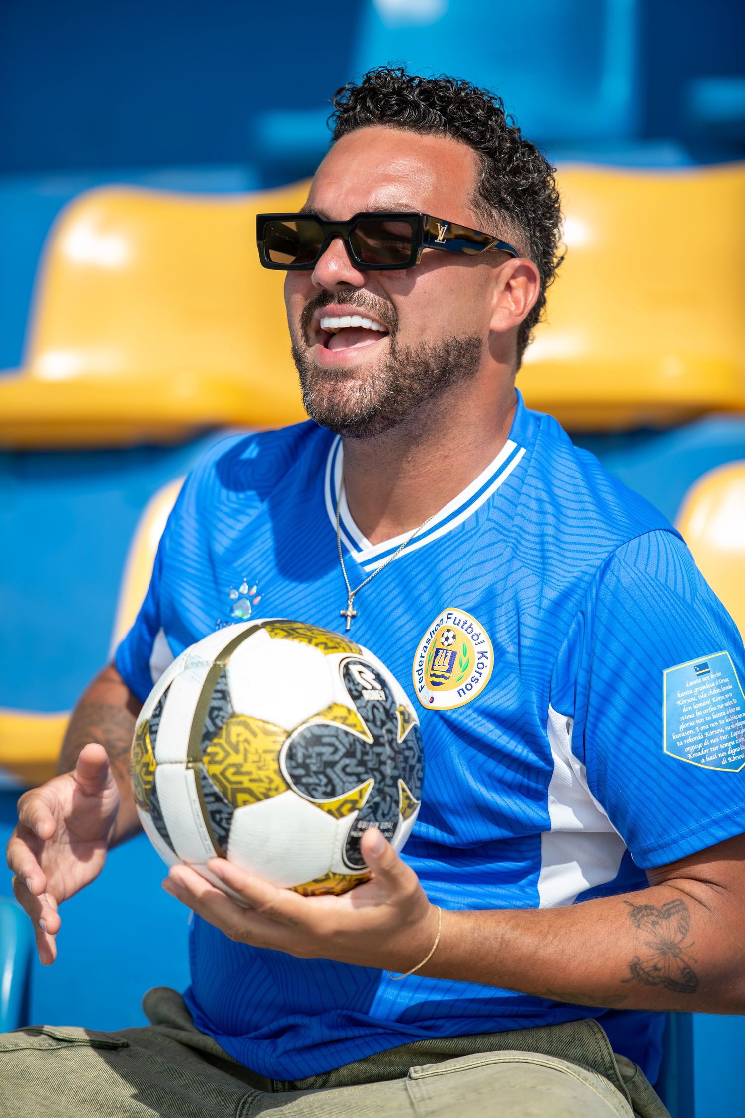 A smiling man in a blue soccer jersey and sunglasses holds a soccer ball while sitting in stadium seats.