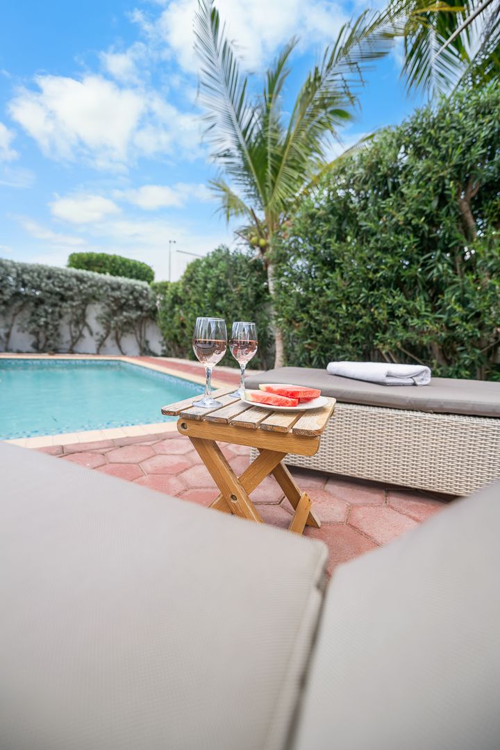 Poolside lounge chairs with a wooden table holding two wine glasses and watermelon slices, surrounded by tropical palms.