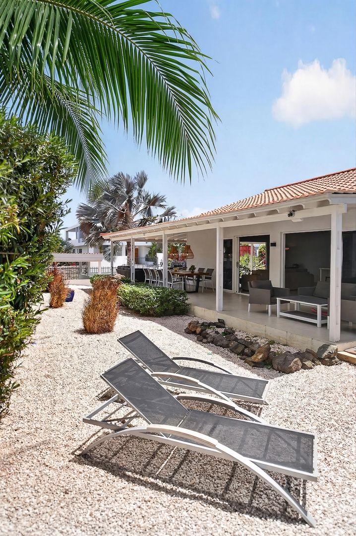 Three lounge chairs on gravel patio beside tropical palm trees and white villa with covered terrace under blue sky.