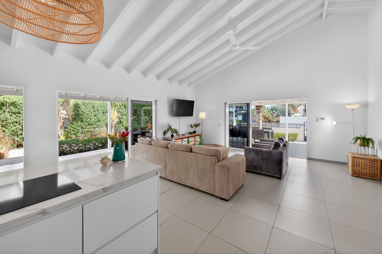 Bright open-plan living room with white beamed ceilings, tile floors, neutral sofas, and large windows overlooking lush greenery.