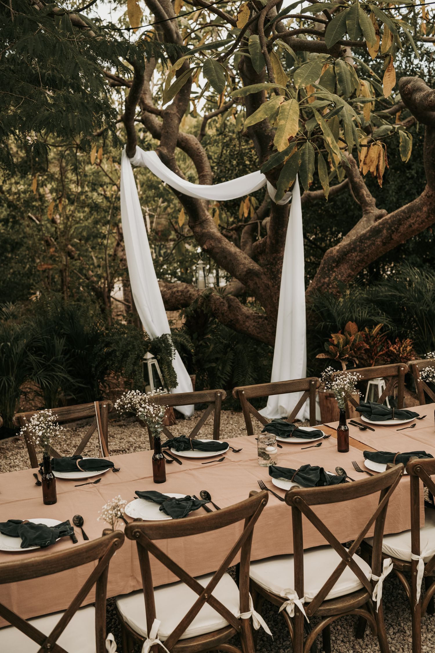 Rustic outdoor wedding setup with white draping, cross-back chairs and table settings under a magnolia tree.