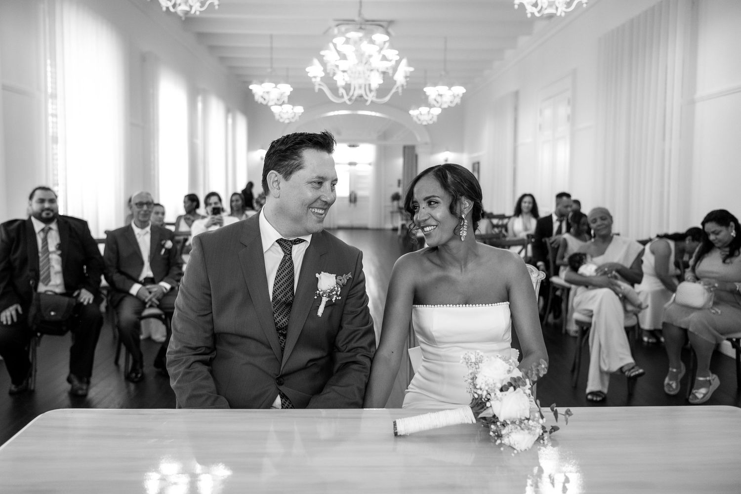 A couple shares a joyful moment during their wedding ceremony in an elegant white room with chandeliers.