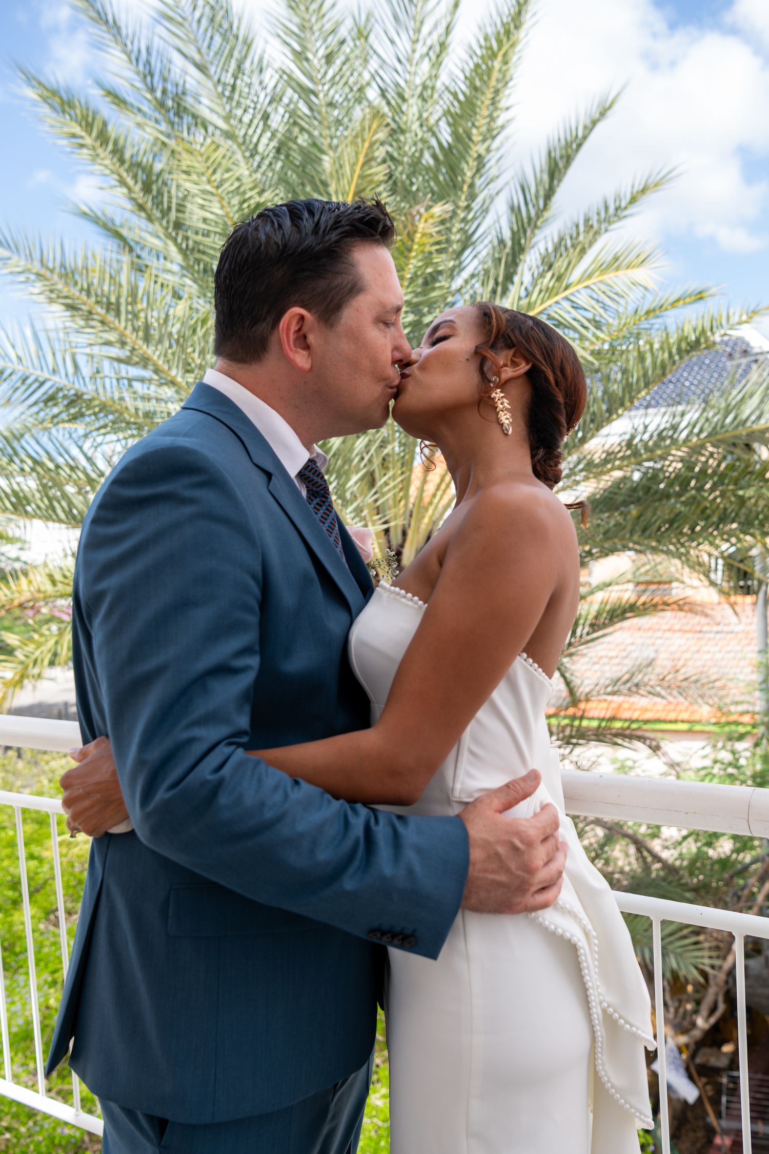 A couple in formal wedding attire share a romantic kiss outdoors with palm trees in the background.