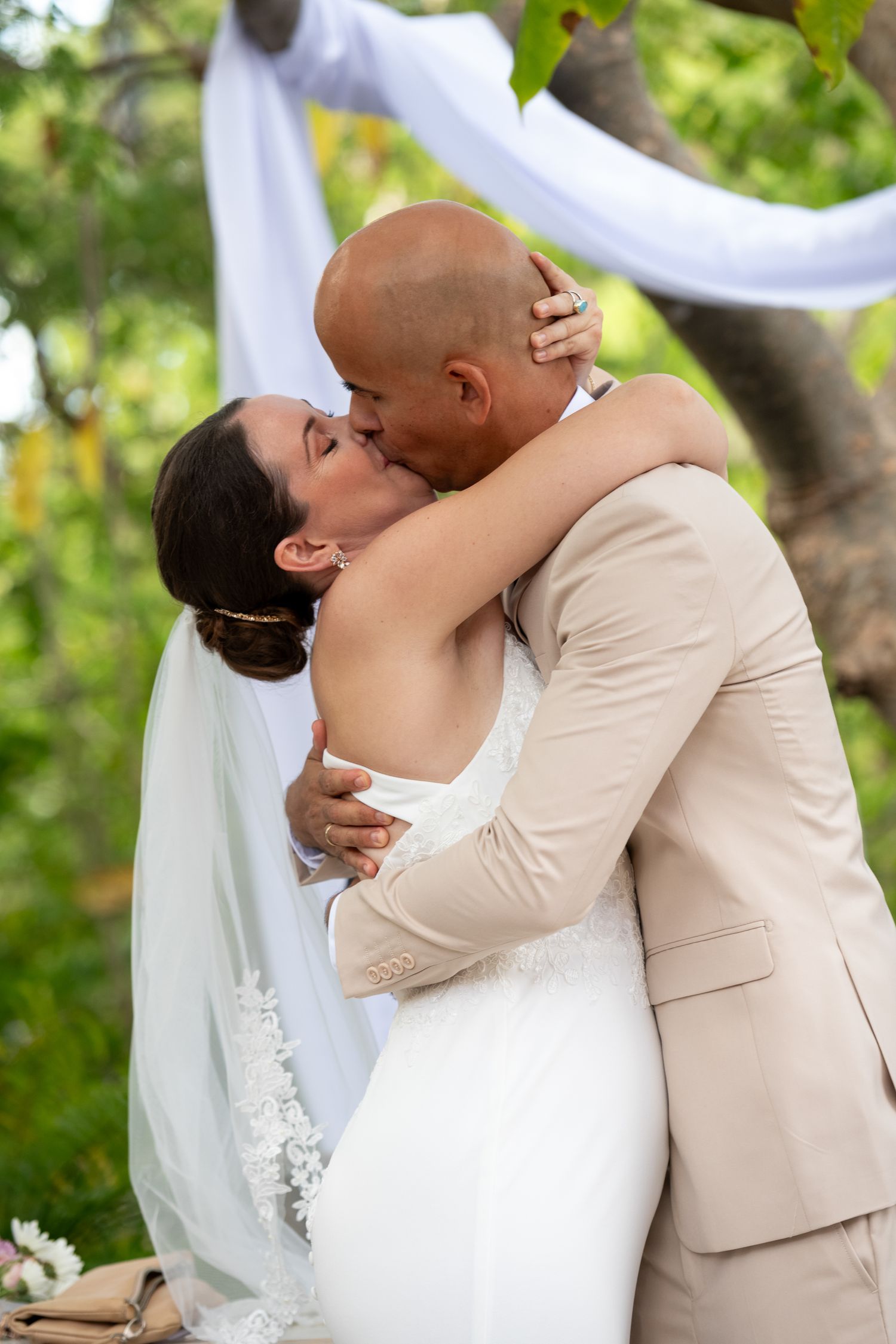 A romantic wedding kiss between newlyweds, with the bride in white and groom in beige, under flowing white drapery outdoors.