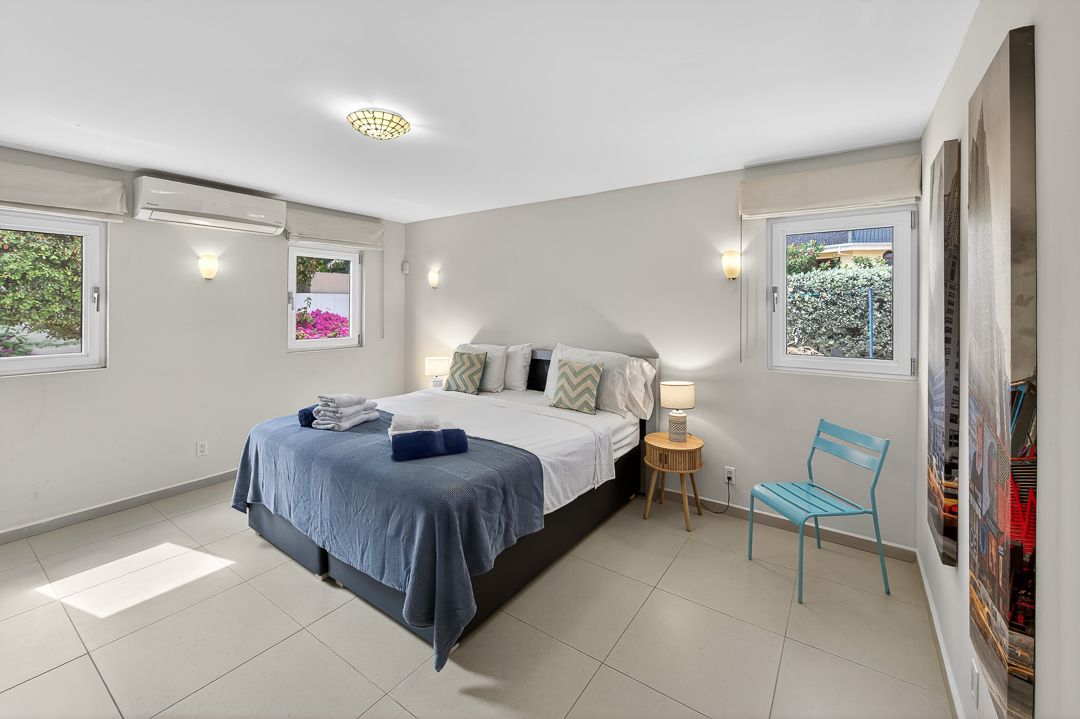 Bright modern bedroom with a king bed, blue linens, teal chair, white walls, tile floors, and two windows with garden views.