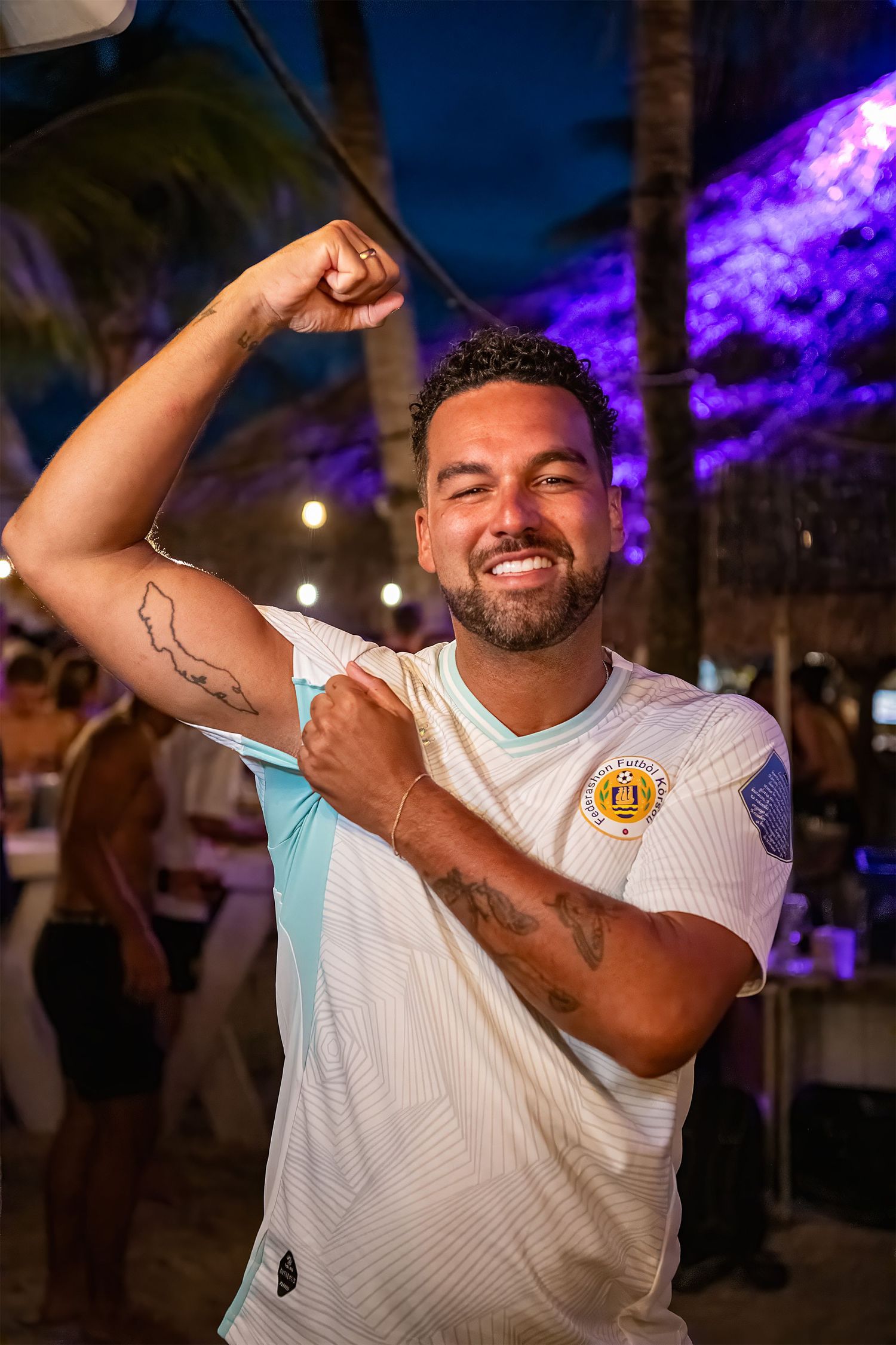 Smiling man flexing his arm tattoo at a tropical outdoor party with purple-lit palm trees at night.