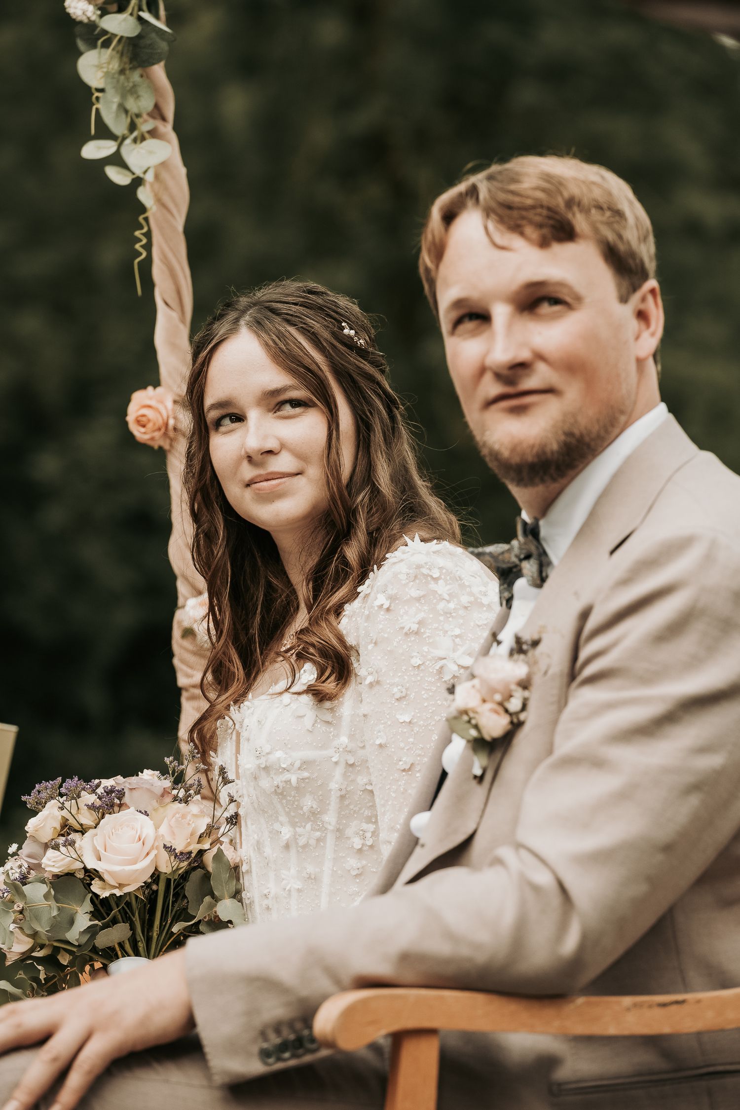 A romantic wedding couple sitting together outdoors in a garden setting with soft lighting and floral bouquet details.