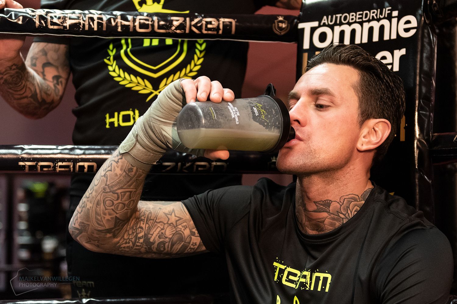 A person wearing a black shirt takes a drink from a bottle in a dimly lit setting.