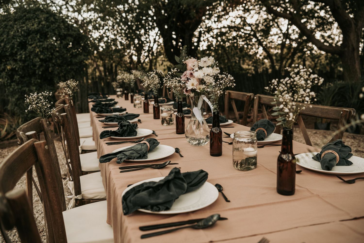 Elegant rustic outdoor table settings with dark plates and wine bottles arranged under trees at dusk.