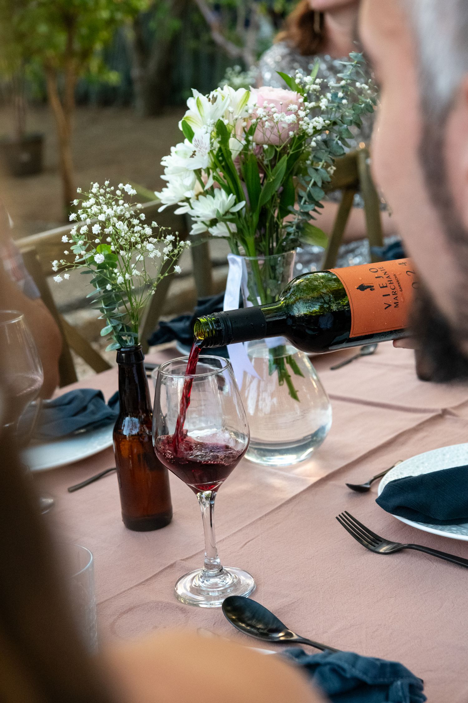 An elegant table setting with red wine being poured, fresh flowers in vases, and navy blue napkins on a blush tablecloth.