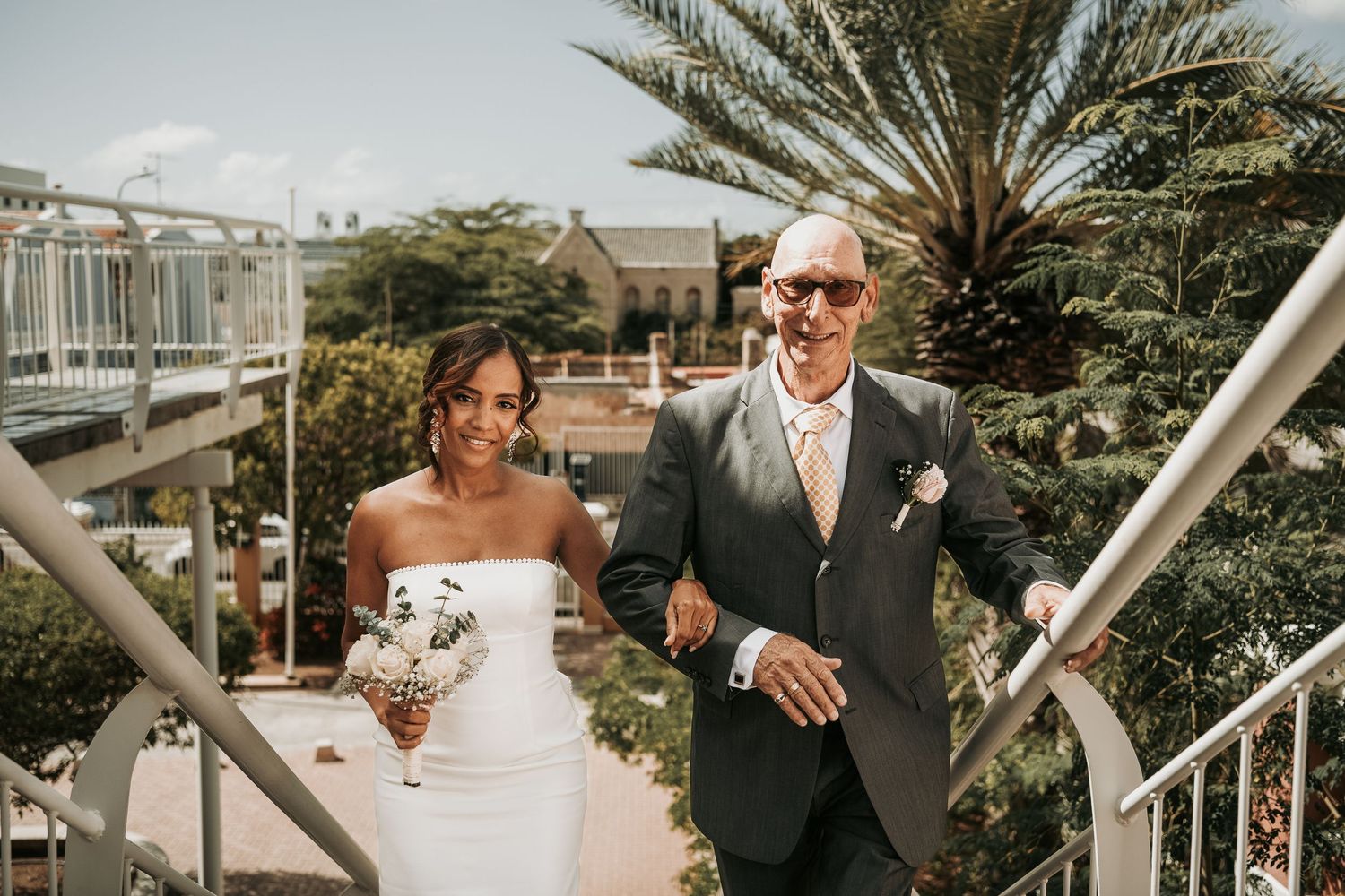 A couple walks down outdoor stairs at a tropical venue during their wedding ceremony.