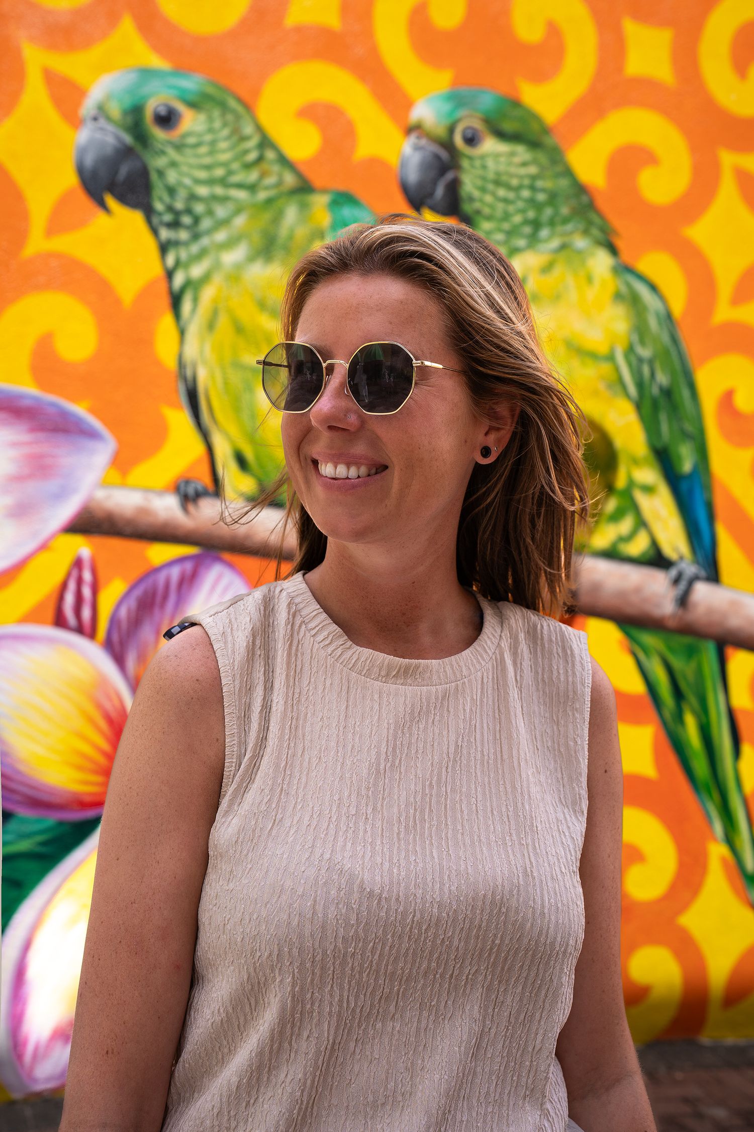 Smiling woman in sunglasses posing before a vibrant mural featuring green parrots on an orange background.