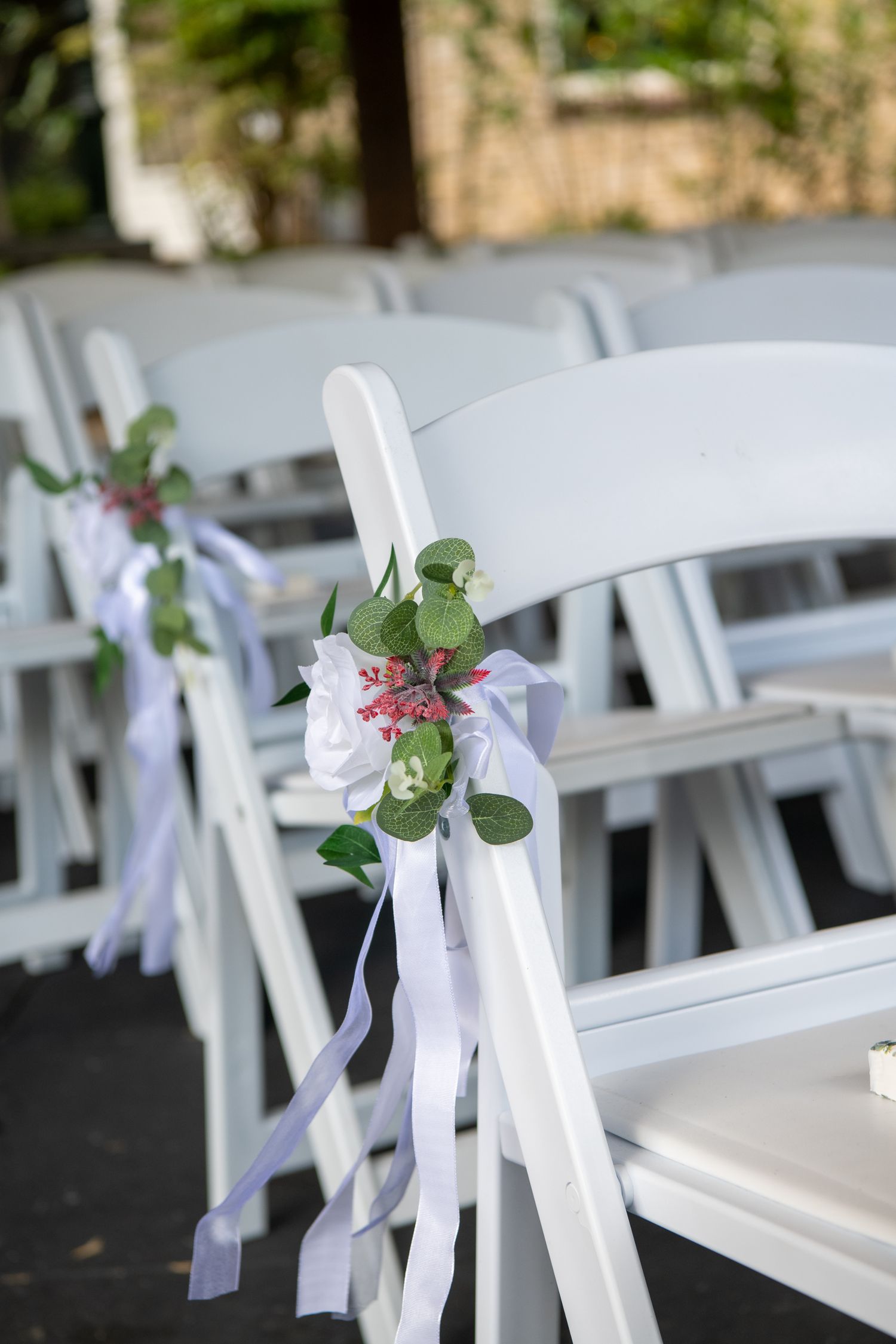 White folding chairs decorated with purple ribbons and pink floral arrangements line up for an outdoor wedding ceremony.