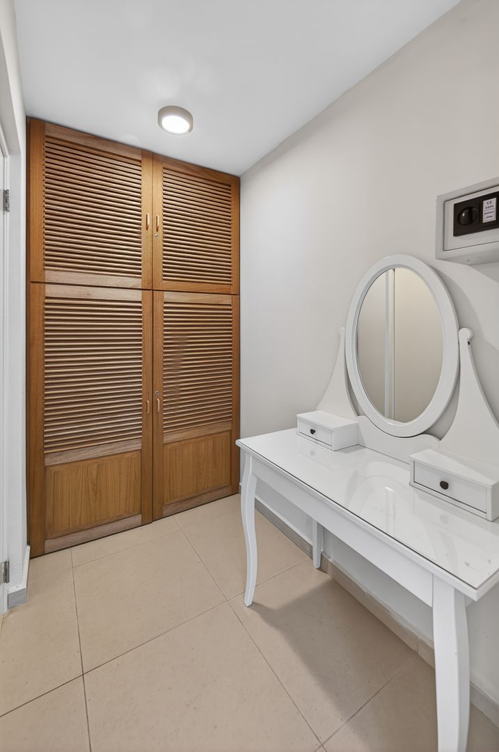 White vanity table with oval mirror beside wooden louvered wardrobe doors on beige tile floor in modern bedroom.