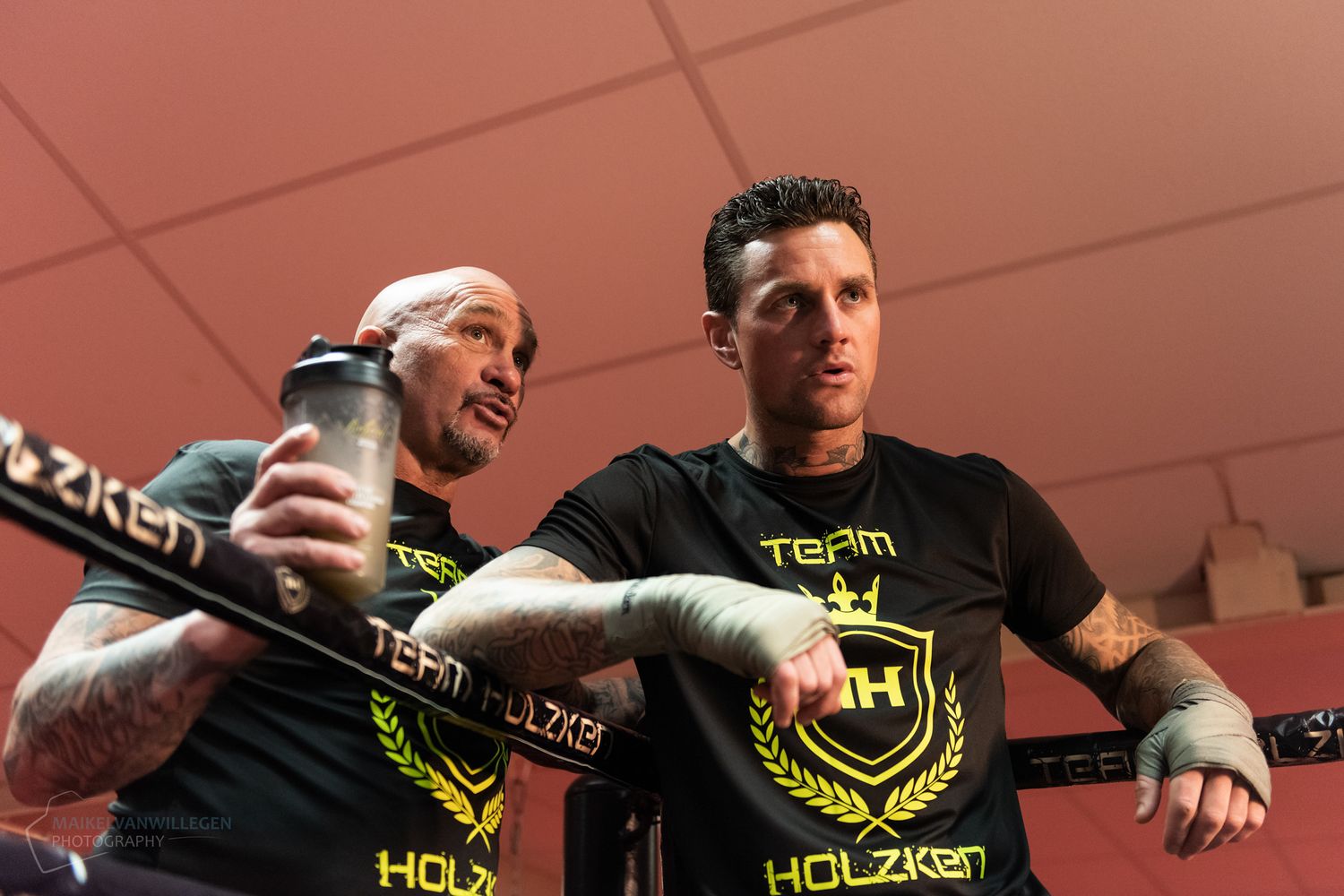Two athletes in black t-shirts with neon green logos train together in an indoor gym setting.