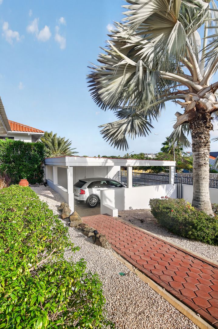 Tropical villa carport with white walls, red brick driveway, parked car, lush palm trees, and manicured shrubs under blue sky.
