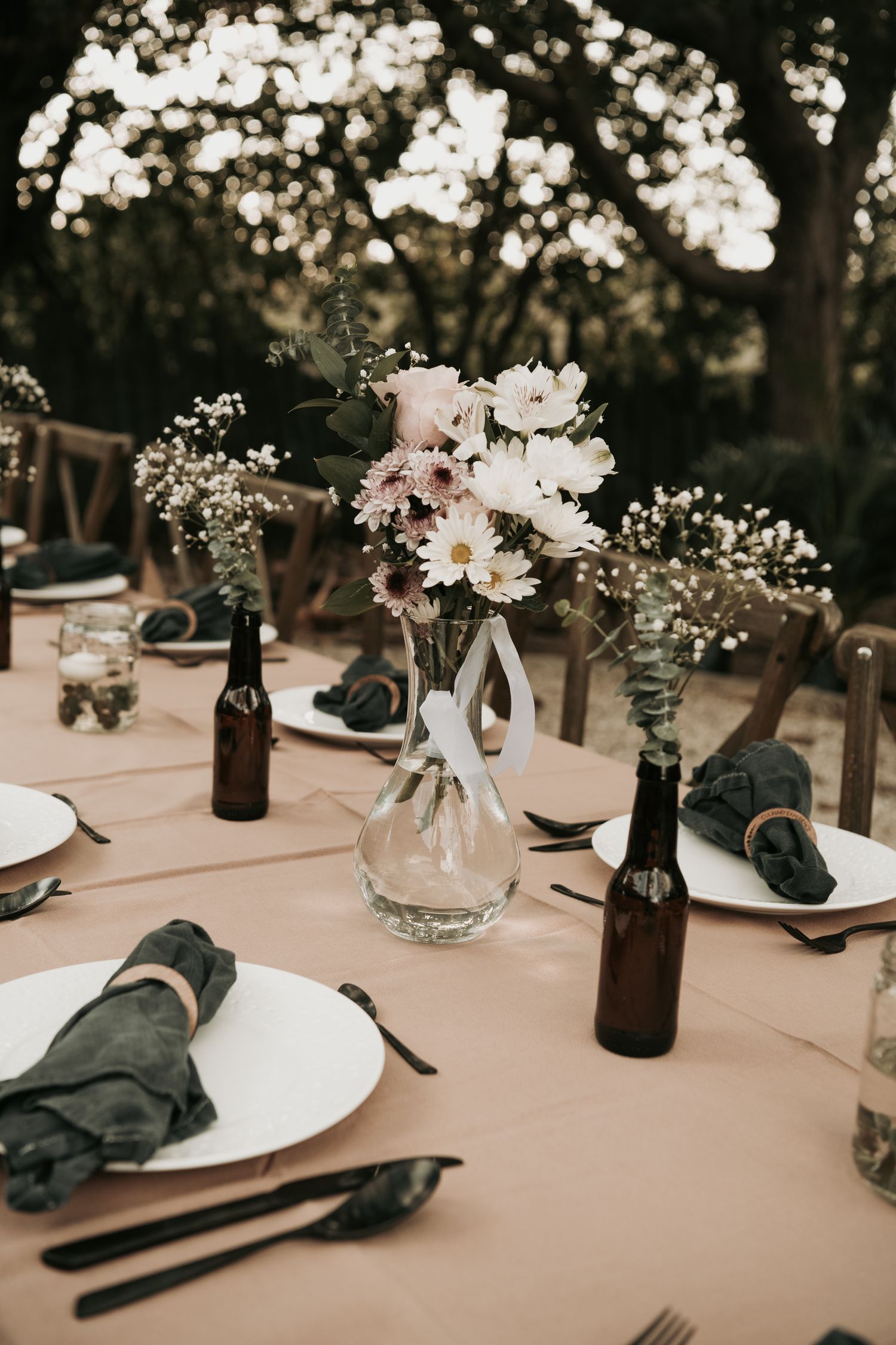 Elegant table settings with brown bottles, white flowers in glass vases, and black napkins create a rustic wedding reception.