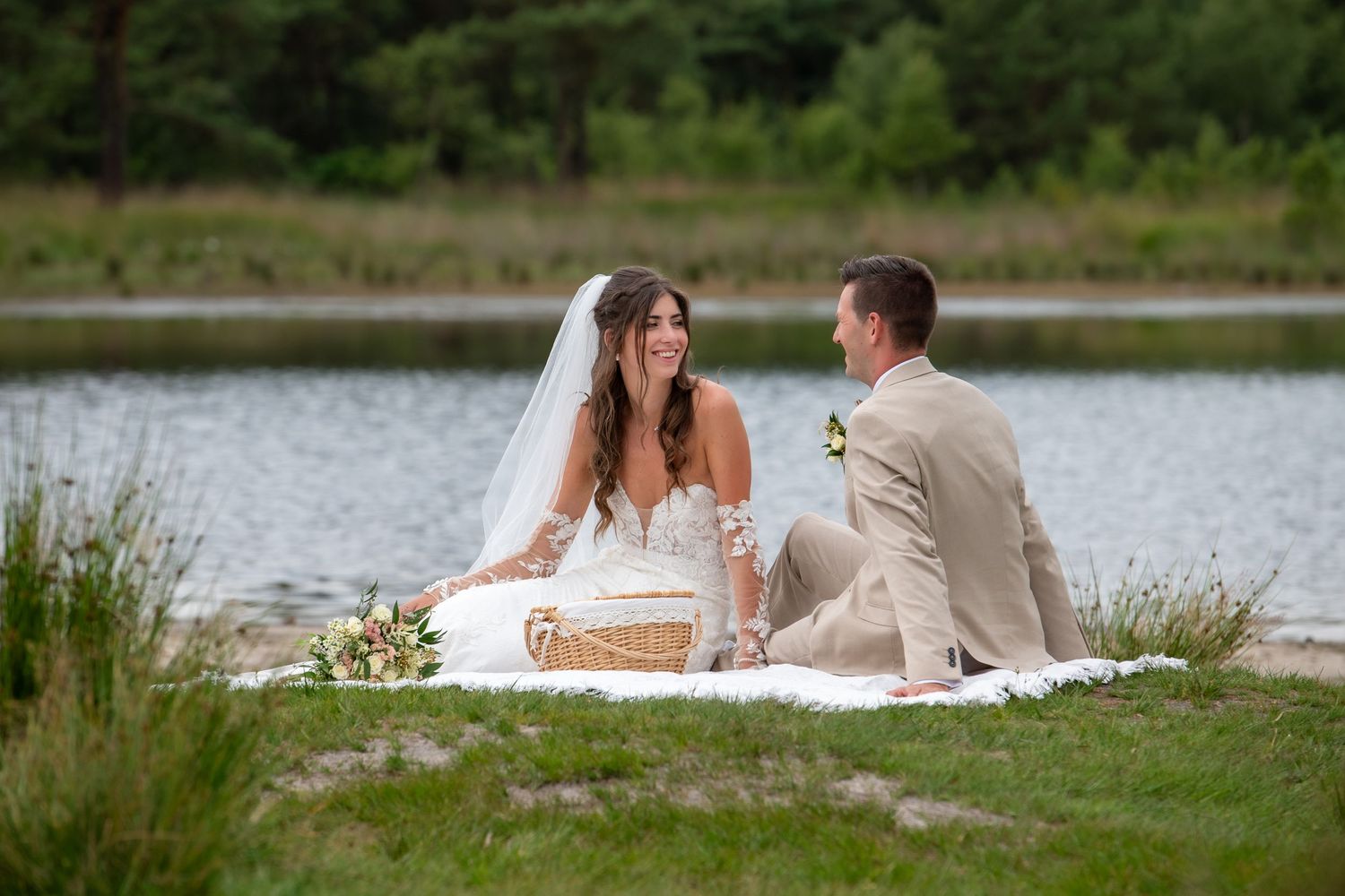 A bride and groom share a moment while sitting on a blanket by a serene lake.