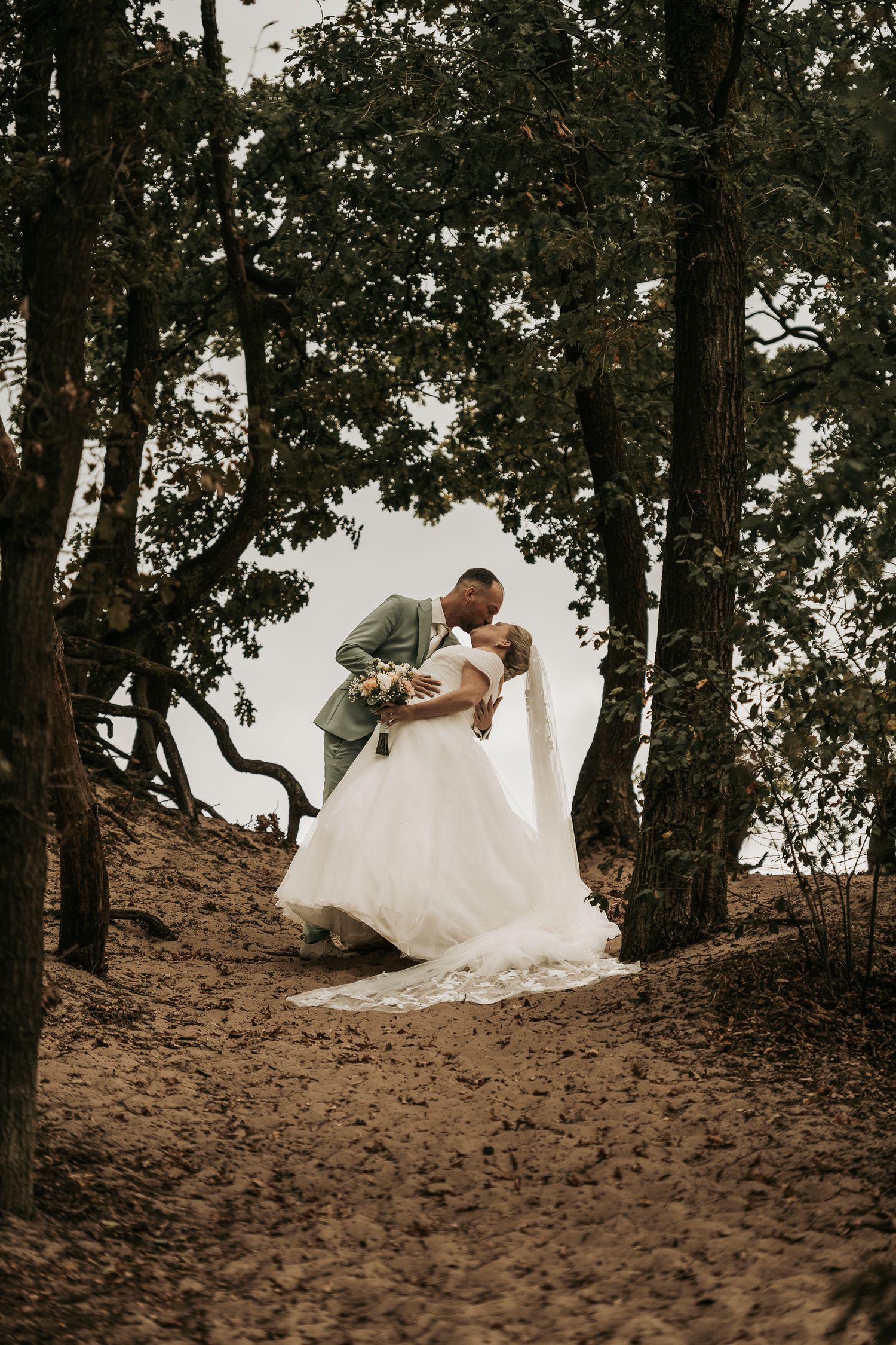 A couple in wedding attire shares a romantic moment on a sandy hillside surrounded by trees.