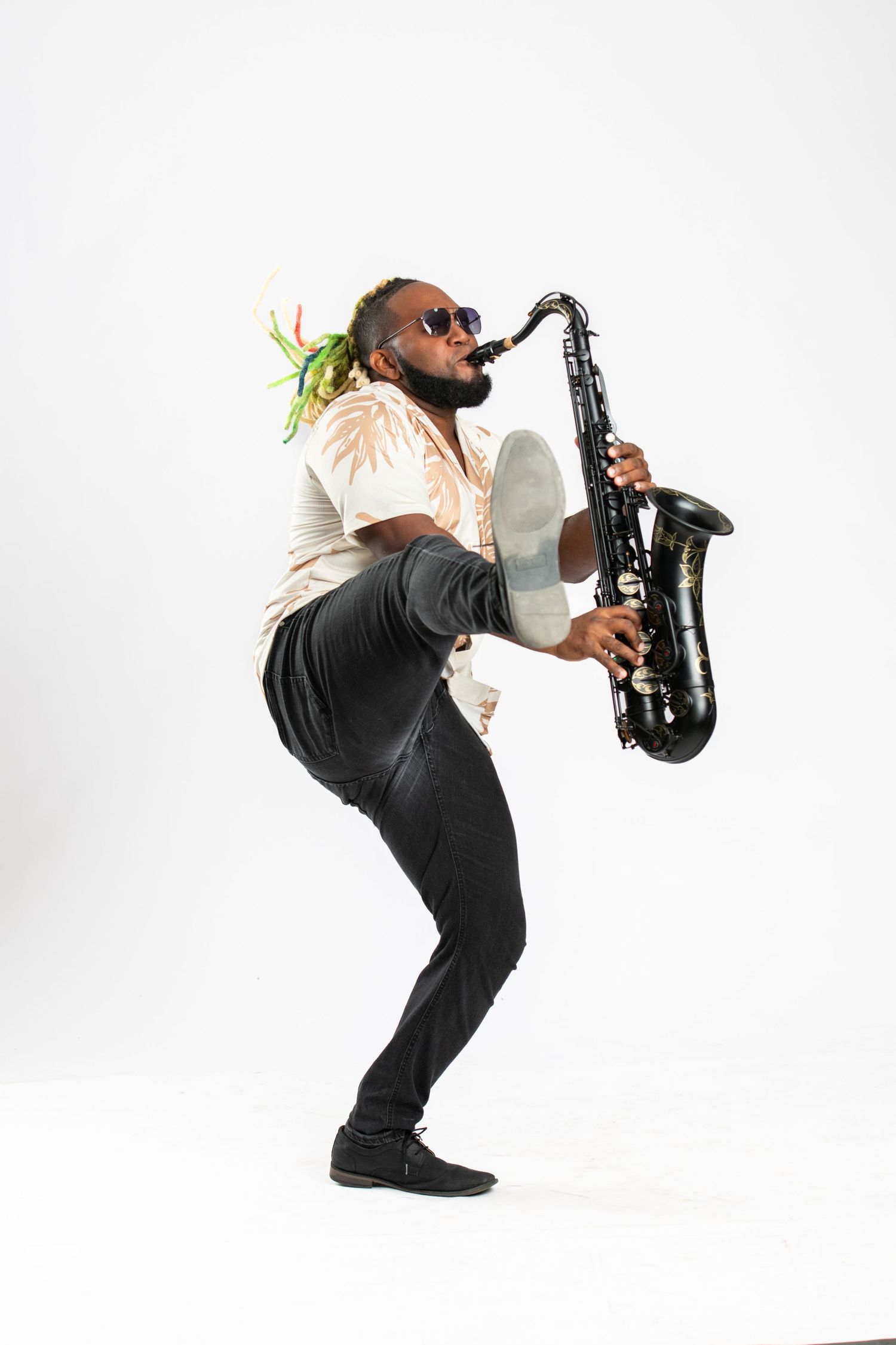 A Black musician with colorful dreadlocks plays a black saxophone energetically, wearing sunglasses and black pants on white background.